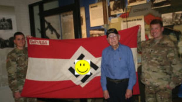  World War II Army veteran Charlie Brown of Olean, New York displays his captured Hitler Youth battle flag from his combat experience in Europe as part of the 258th Field Artillery Battalion with Lt. Col. Peter Mehling, right, and Capt. Steven Kerr, left, the current battalion commander and operations officer of the New York Army National Guard’s 1st Battalion, 258th Field Artillery at the New York State Military Museum in Saratoga Springs, New York November 20, 2017. ( U.S. National Guard photo by Col. Richard Goldenberg.)