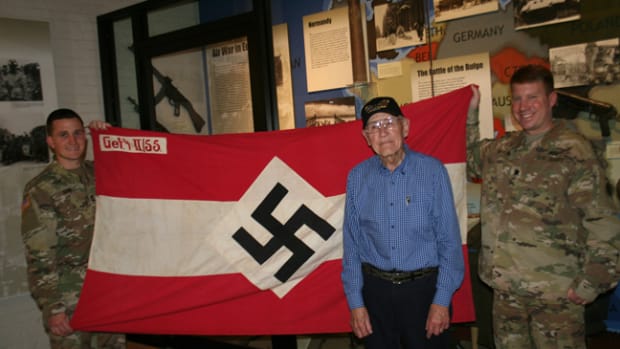  World War II Army veteran Charlie Brown of Olean, New York displays his captured Hitler Youth battle flag from his combat experience in Europe as part of the 258th Field Artillery Battalion with Lt. Col. Peter Mehling, right, and Capt. Steven Kerr, left, the current battalion commander and operations officer of the New York Army National Guard’s 1st Battalion, 258th Field Artillery at the New York State Military Museum in Saratoga Springs, New York November 20, 2017. ( U.S. National Guard photo by Col. Richard Goldenberg.)