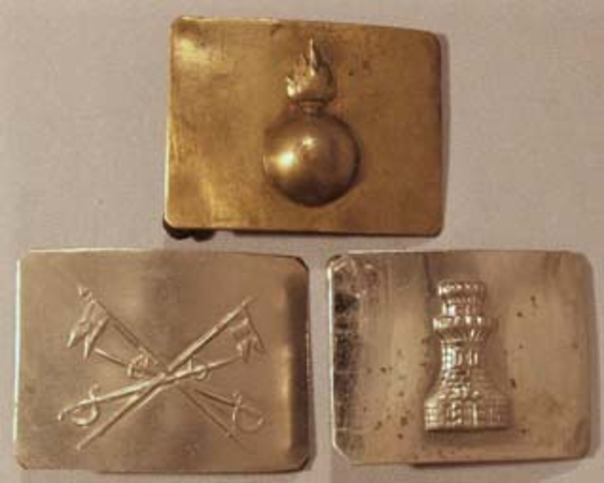 Combat Services Belt Plates. A flaming bomb emblem represents the Artillery on the brass plate at the top. The traditional weapons of a horseman, sabers and lances, are cross on the nickel plate used by the Spanish Cavalry, shown bottom left. On the lower right the nickel finished plate with a castle tower was worn by Engineers.