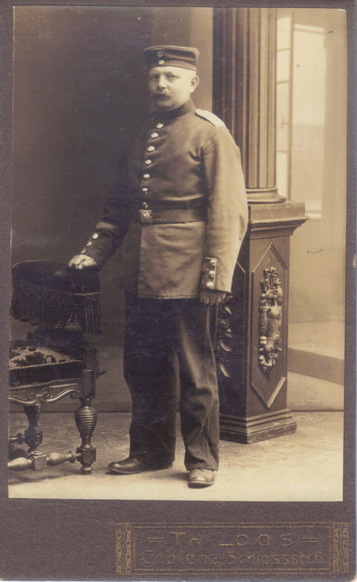 Another non-US soldier portrait, this time a German soldier poses with the chair and the open-window backdrop. Note that in this photograph there is also an ornate column that appears to have a hunting motif-the most visible symbol on it appears to be a dead rabbit in addition to all of the now-familiar Loos props: chair, ornate column, and open-window backdrop.