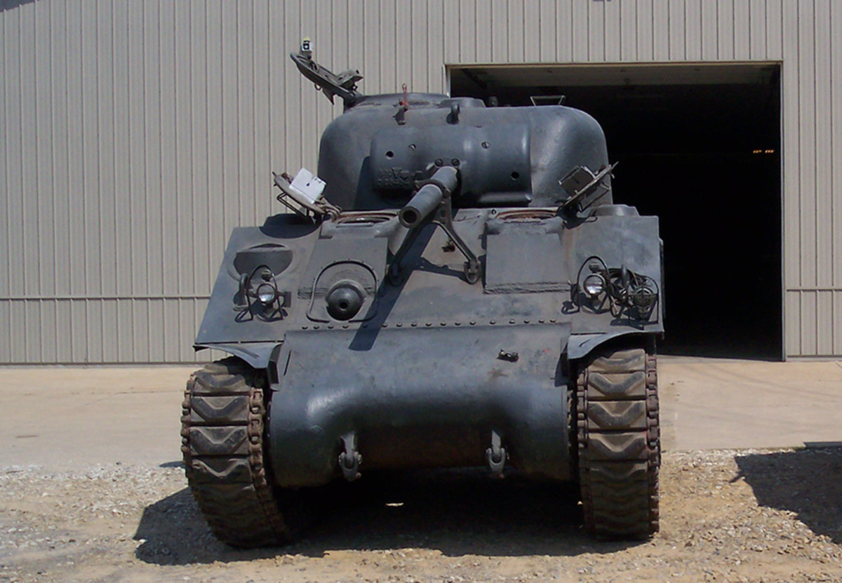 Modern photo, head on, of an unrestored M4A3 in front of a steel building.