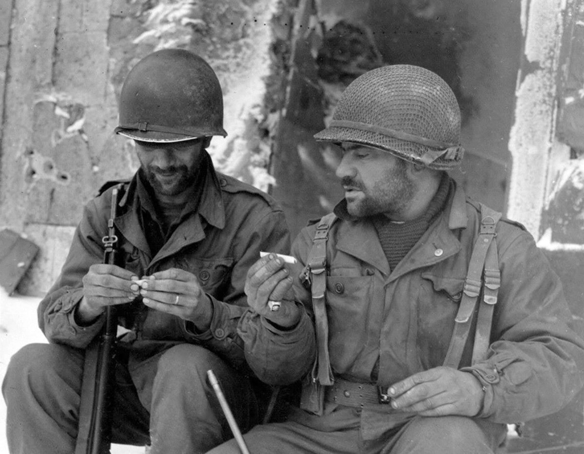 WWII photo of two American GIs rolling cigarettes. Both wear M1 helmets, one has net cover.
