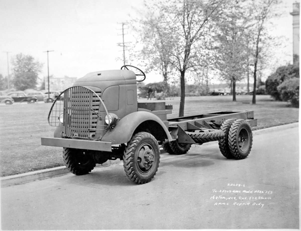 A GMC AFKX-352 1 1/2-3-ton 4x4 cab-over-engine chassis for a small-arms repair truck is seen from the front left, showing the shape of the brush guard, the location of the spare tire and its carrier, and the presence of dual rear wheels. A strut on each side of the central part of the brush guard was attached to the clip, to the outer sides of the radiator louvers. The rear of the left strut is next to the left service headlight. A blackout lamp, in a small, bullet-shaped housing, was on the outboard side of each service-headlight assembly
