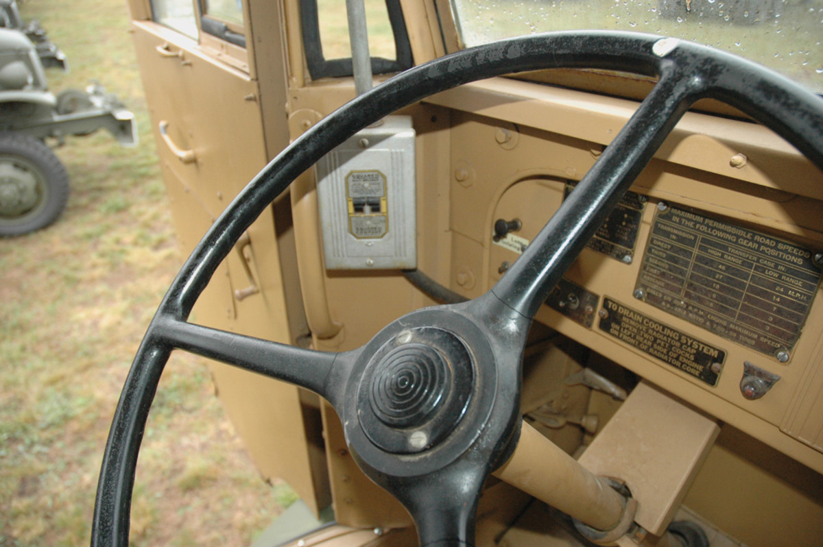 A Square D multi-breaker, with an electrical cable running from below it forward to the electrical box on the exterior of the left side of the cowl, and a conduit passing from the top of the box up to the ceiling, providing power for the electrical lights and receptacles. Visible is the steering wheel, the bracket for the steering column, and data plates with instructions on draining the cooling system and maximum possible road speeds in different transmission and transfer-case gear positions.
