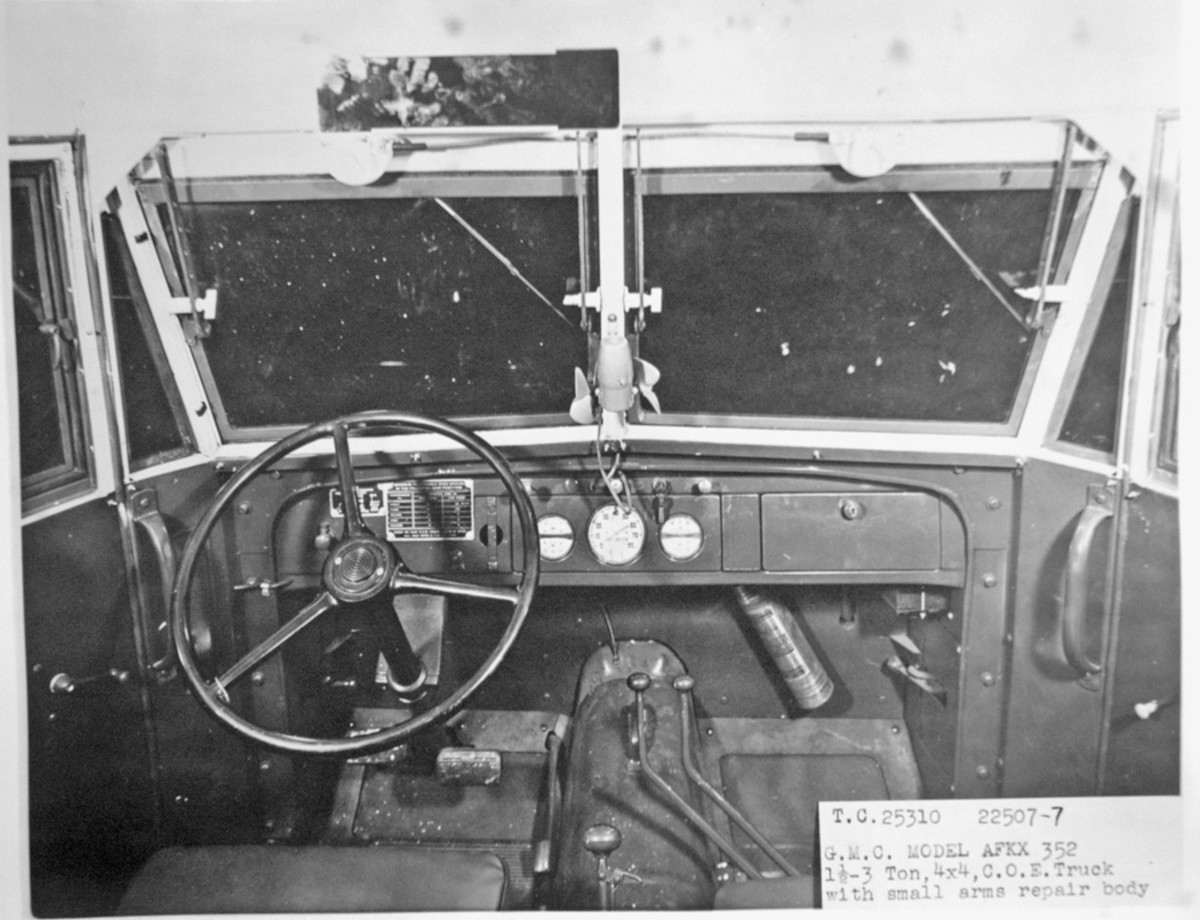 The interior of the cab of an AFKX-352 is on view; the same photograph was used as an illustration in the maintenance manual for this series of trucks, TM 10-1401. On the dash are data plates (left), instruments (center), and package compartment (or glove compartment; right), below which is a fire extinguisher. Flanking the steering column are the clutch (left) and brake (right) pedals, with the small pedal for the accelerator on a long lever to the left of the tunnel. At the lower center is the transmission shift lever, to the front of which are the transfer-case speed shift lever (left) and the front-axle declutching lever (right).