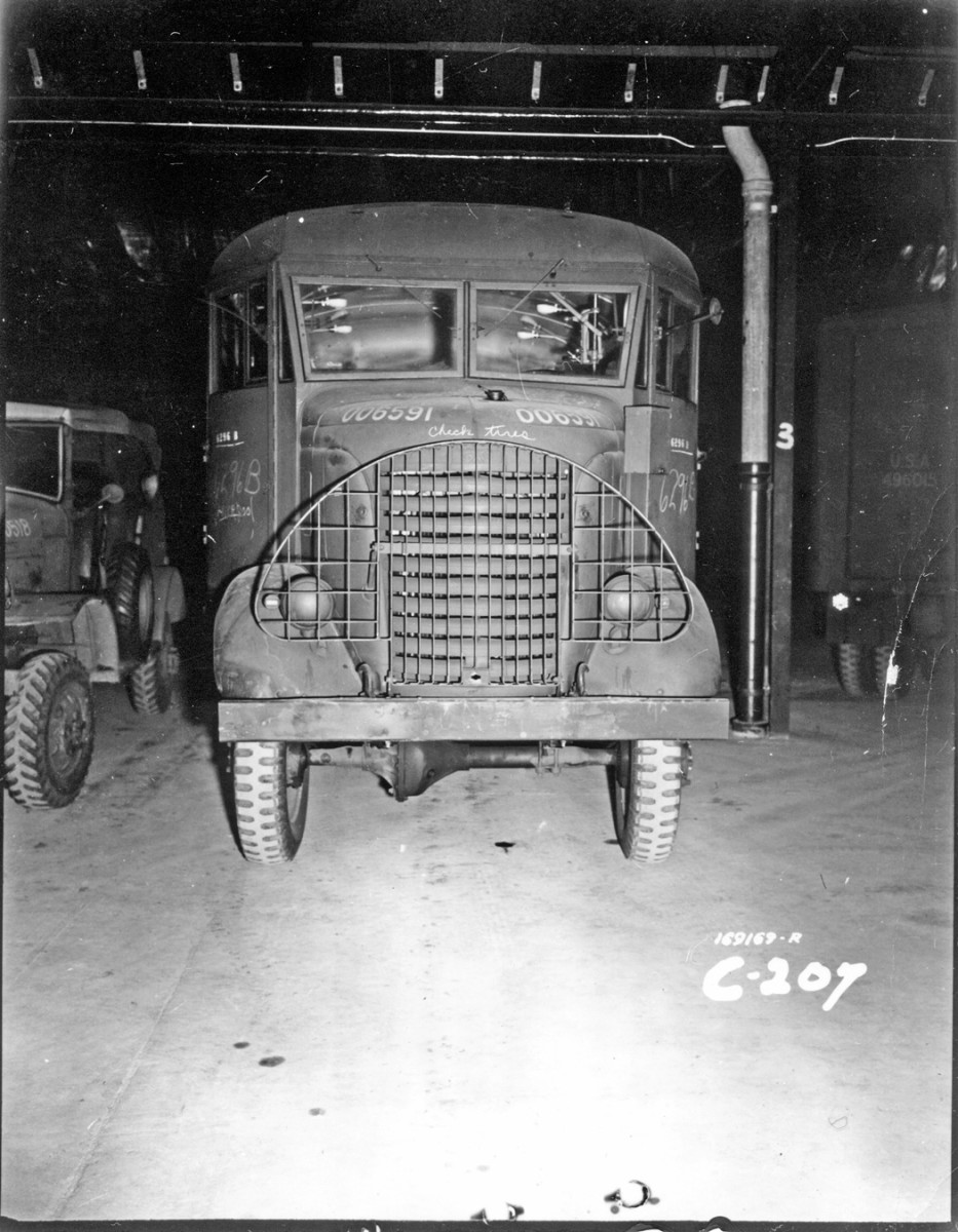 A GMC AFKX-352 small-arms repair truck, registration number 006591, is being prepared for service in a shop at the U.S. Army Ordnance depot at Ashchurch, England, on 5 October 1942. An electrical box is mounted on the left side of the cowl. “Check tires” is chalked above the radiator brush guard, while “6296 B” is stenciled in white and also marked in chalk on both cab doors. What appears to be “Liverpool” is chalked near the bottom of the right door