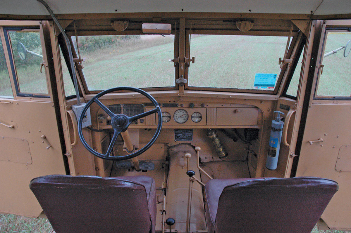 In a view of the cab, between the seats are, left to right, the hand-brake lever, the power takeoff control, and the transmission shift lever. To the left of the steering wheel is a Square D multi-breaker, with an electrical cable running from below it forward to the electrical box on the exterior of the left side of the cowl, and a conduit passing from the top of the box up to the ceiling, providing power for the electrical lights and receptacles.