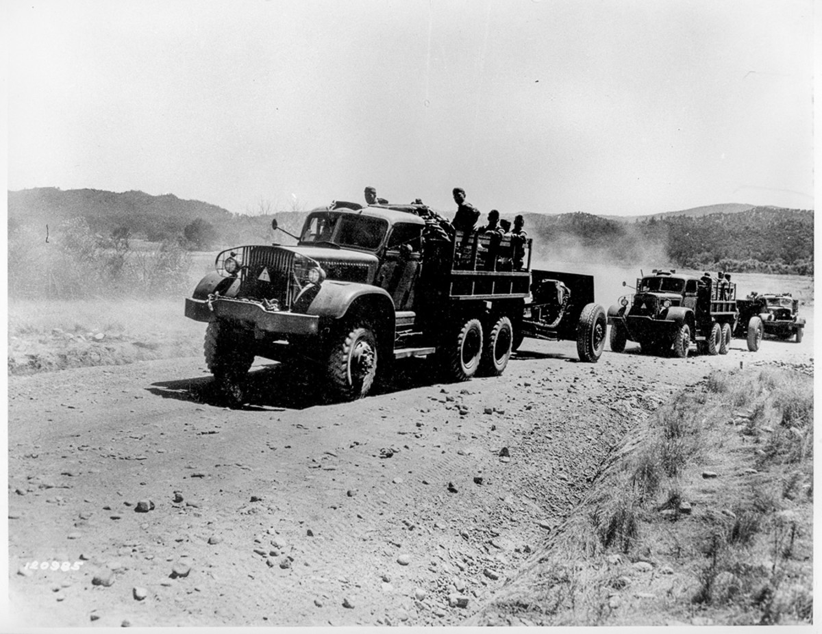 Three Diamond T Model 967s are serving as prime movers for 155mm Howitzers M1917 or M1918—the role for which they initially were intended—during a field exercise. A small placard, a light-colored triangle with a cross on the center, is on the brush guard of each truck. Pioneer tool racks are above the left running boards.