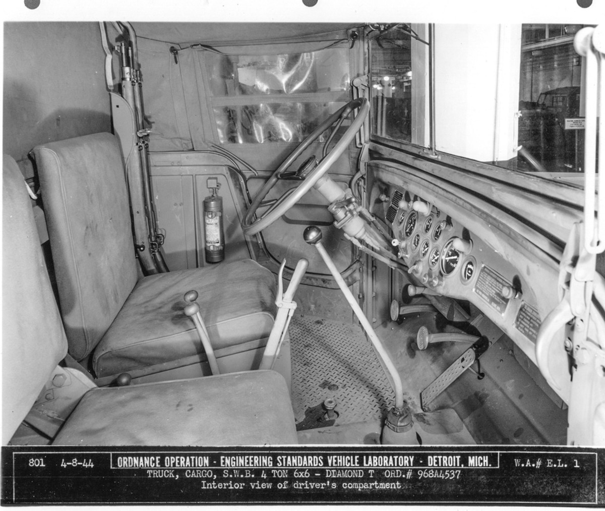 An 8 April 1944 photo depicts the interior of the open cab of Diamond T serial number 968A4537. Between the seats are, rear to front, the power takeoff shift lever (only the knob is visible), the transfer shift lever and transfer declutch lever (side by side), the parking-brake lever, and the transmission shift lever. A fire extinguisher is on the far side of the cab, to the front of the rifle brackets.