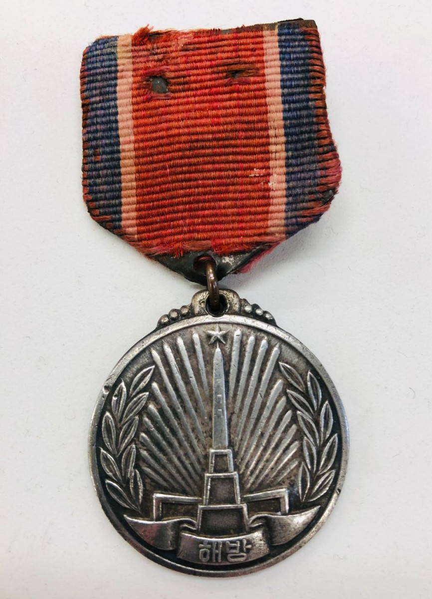 The Korean Liberation Commemoration Medal is a 33mm silver medal with the Liberation Commemoration Tower embossed on a rayed background. Laurel leaves are on each side and the word “Liberation” on the ribbon at the bottom in two characters.
