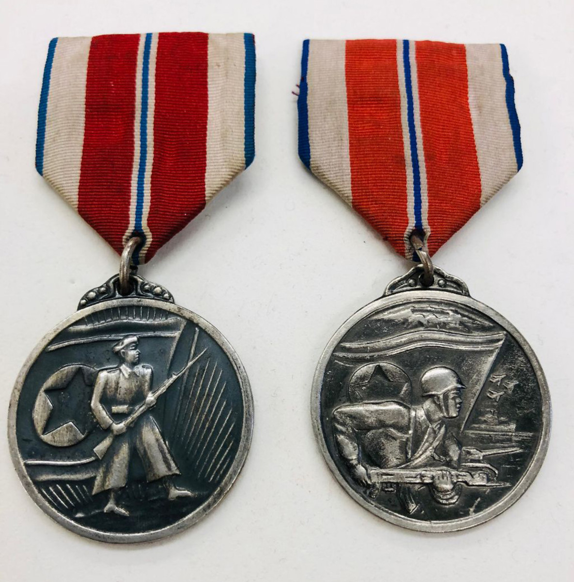 The first version of the Military Merit Medal shows a North Korean soldier wearing an overcoat and carrying a rifle with a large North Korean Flag behind him. The current version shows the upper body of a North Korean soldier carrying an AK-47 rifle. A partial North Korean Flag with a ship and two aircraft are found to the right.