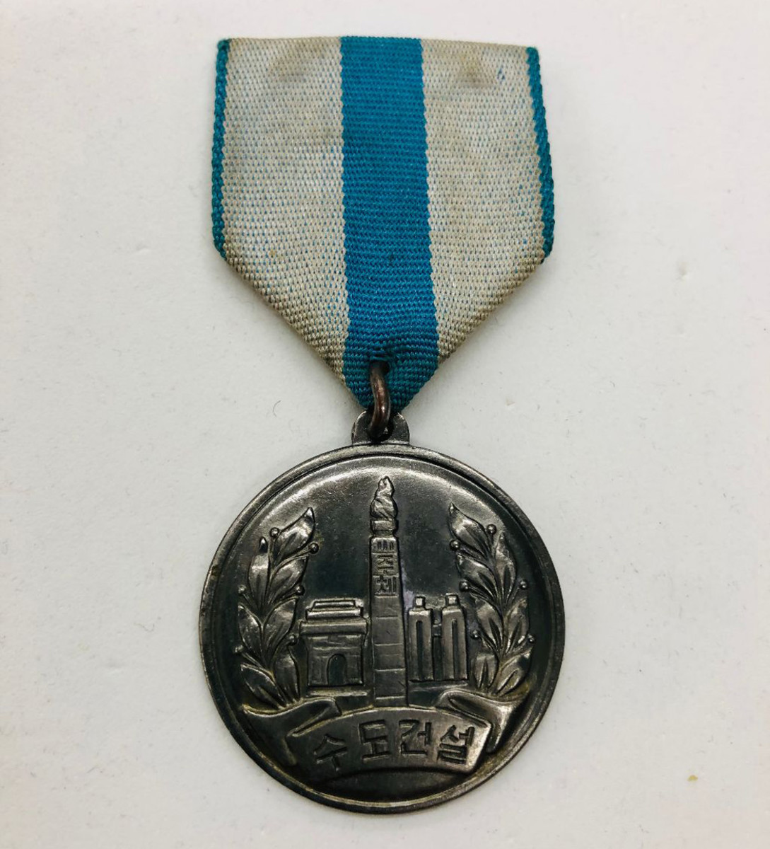 The 32mm silver Capital City Construction Medal shows the Juche tower in the center and the Arch of Triumph and two towers of the Koryo Hotel left and right. The Korean characters spell Pyongyang.